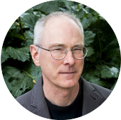 A person wearing glasses and a dark jacket stands among a bush with large green leaves.