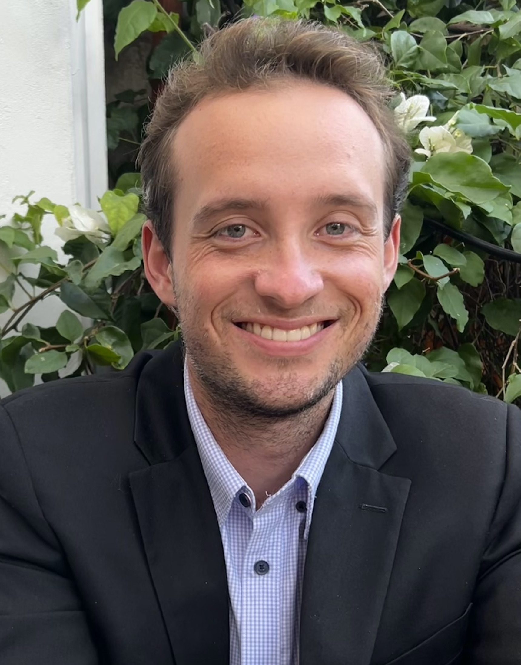 White male, with short, dirty blonde hair, in his twenties wearing a black and blue suit in front of green ivy.suit